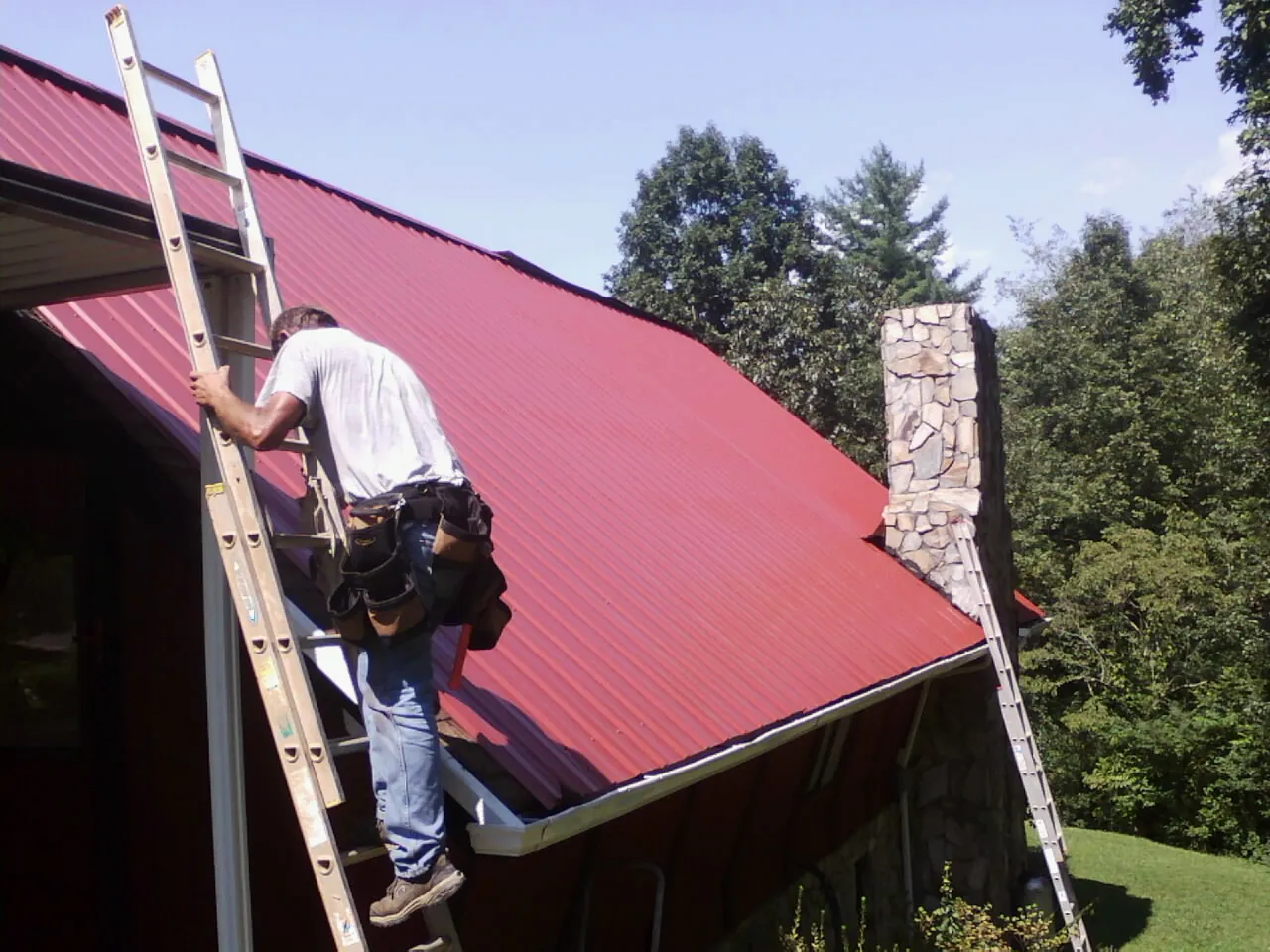 Roofer on a ladder beside a red metal roof and stone chimney — Soffit & Fascia Repair by Roofing Brothers in West Jefferson